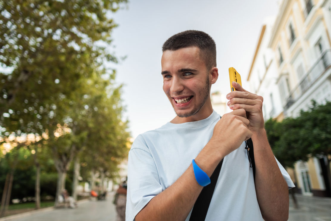 Ein junger blinder Mann hält sein Smartphone am Ohr, während er draussen auf der Strasse unterwegs ist. Der Hintergrund ist unscharf, der Fokus auf ihm liegt. Er nutzt vermutlich die Voice Over-Funktion, um Informationen akustisch zu erhalten und so selbstständig und barrierefrei zu kommunizieren,.